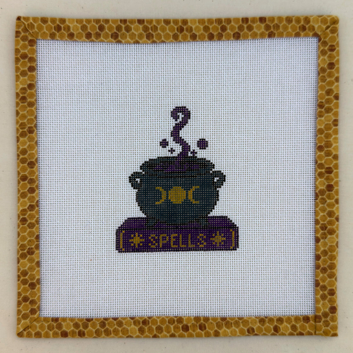 Hand-painted needlepoint canvas of a black cauldron on a purple book marked Spells