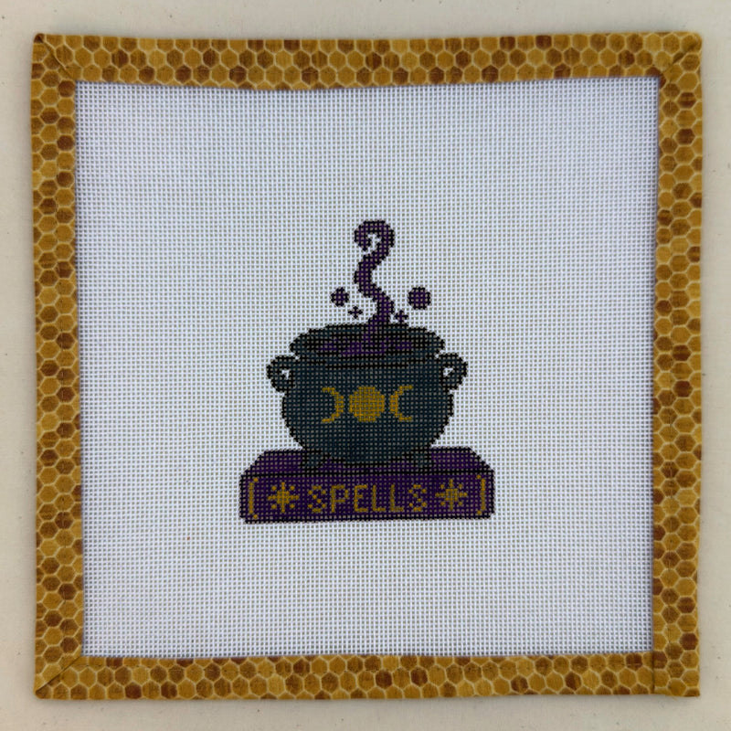 Hand-painted needlepoint canvas of a black cauldron on a purple book marked Spells