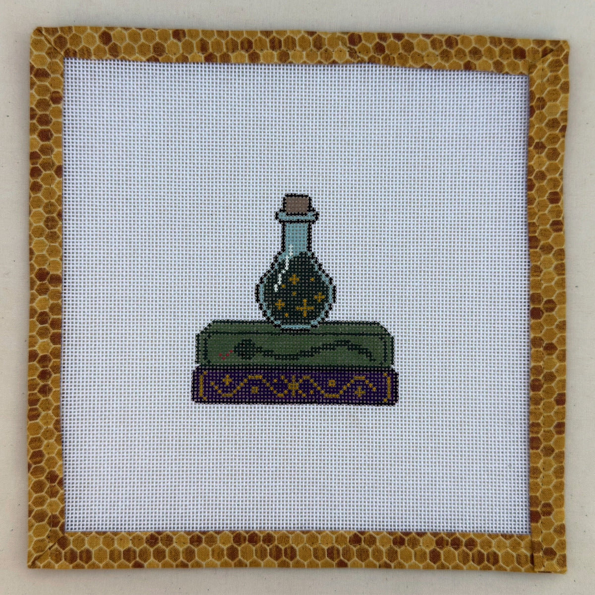 Hand-painted needlepoint canvas of a green potion bottle on stacked spellbooks