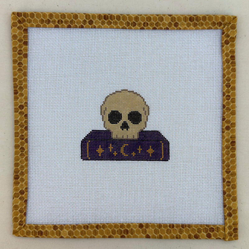 Hand-painted needlepoint canvas of a pale skull on a purple spellbook